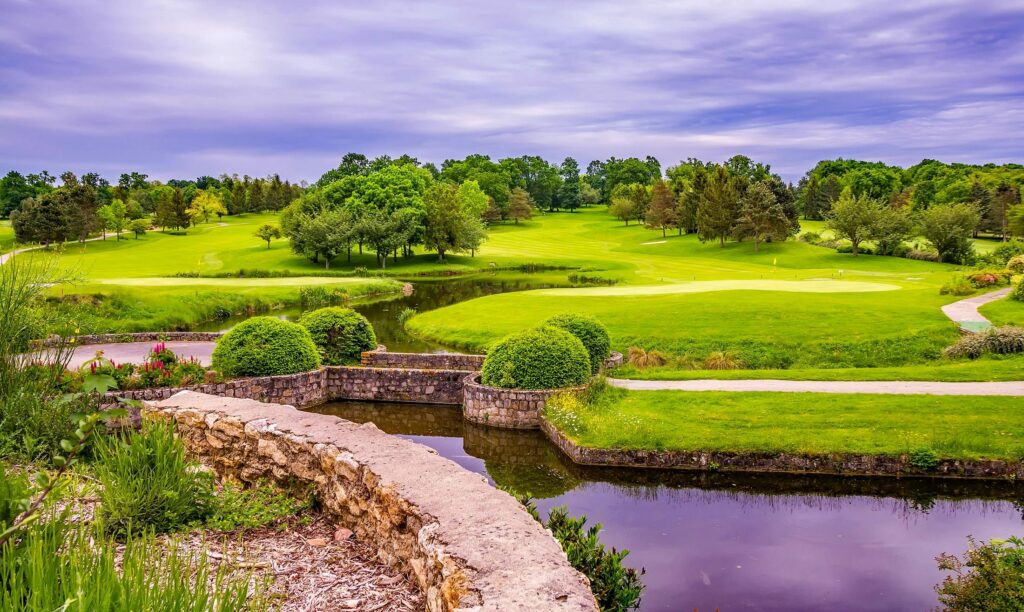 Serene countryside view with lush greenery, pond, and vibrant trees under a cloudy sky.